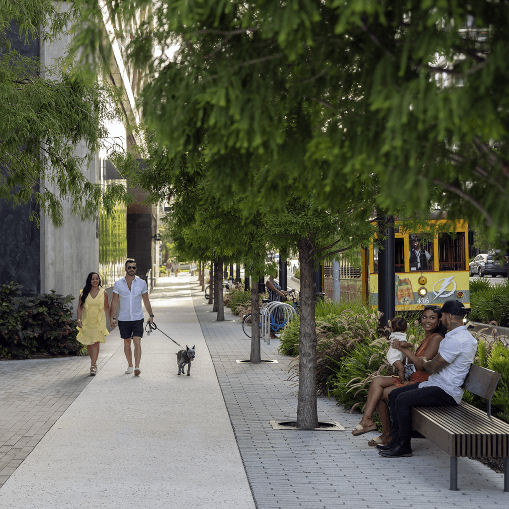 A couple walks their dog while another couple sit on a bench outdoors in Water Street Tampa