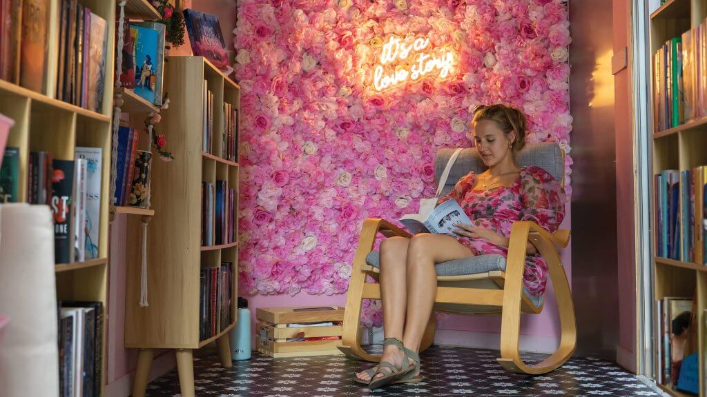 A woman reads a book in a pop-up bookstore on water street under a script neon light that says its's a love story